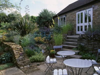 Patio With Metal Chairs And Tables, Stone Wallls Planted With Achilleas, And Phormium by Clive Nichols Pricing Limited Edition Print image