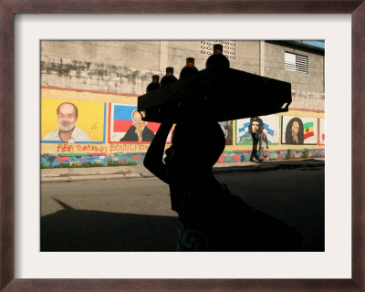A Boy Carrying Bottles On His Head Passes By A Wall With Pictures Of Haitian President Renel Preval by Ariana Cubillos Pricing Limited Edition Print image