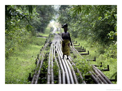 A Woman Walks Along An Oil Pipeline Near Shell's Utorogu Flow Station, Nigeria, January 15, 2006 by George Osodi Pricing Limited Edition Print image