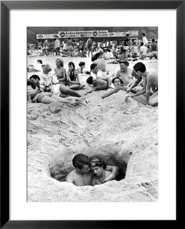 Couple Cuddling While Sitting In A Hole As Others Enjoy The Beach On The 4Th Of July by Ralph Crane Pricing Limited Edition Print image