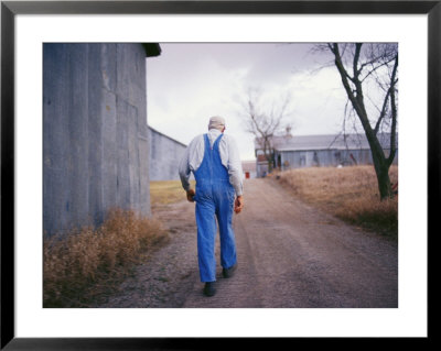 An Elderly Farmer In Overalls Walks Along A Dirt Road Past A Barn by Joel Sartore Pricing Limited Edition Print image