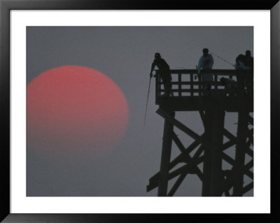 A Harvest Moon Rises Over A Pier Where A Group Of Fishermen Cast Their Lines by Joel Sartore Pricing Limited Edition Print image