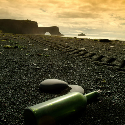 A Green Bottle Lying On A Beach, Cliff Dyrholaey In Background, South Iceland by Larus Karl Ingasson Pricing Limited Edition Print image