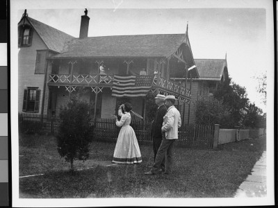 Two Men Watching As A Young Woman Plays Lawn Tennis; Other Women Watch From A Second Story Porch by Wallace G. Levison Pricing Limited Edition Print image