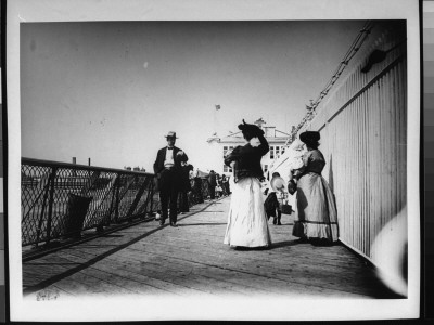 Well-Dressed Men And Women Walking On A Boardwalk Next To A Pier And A Body Of Water by George B. Brainerd Pricing Limited Edition Print image