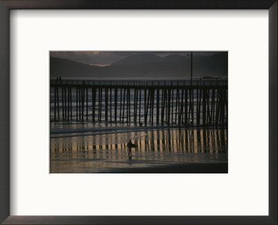 A Surfer Walks Up The Beach Near A Pier At Twilight by Michael S. Lewis Pricing Limited Edition Print image