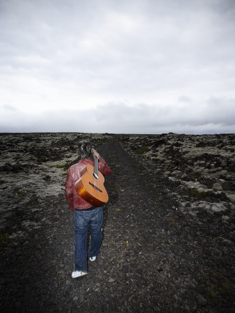 A Teenage Boy Walking With An Acoustic Guitar On A Gravel Road In Iceland by Atli Mar Pricing Limited Edition Print image