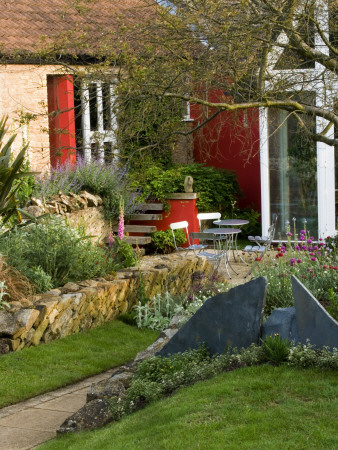 View Across The Lawn To The Patio With Metal Table And Chairs, Red Wall And Angled Drystone Wall by Clive Nichols Pricing Limited Edition Print image